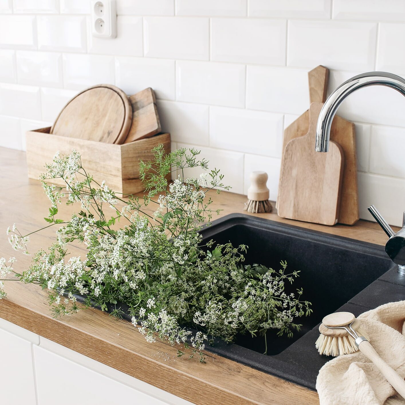 Closeup of kitchen interior. White brick wall, metro tiles, wooden countertops with chopping boards. Cow parsley plants in black sink. Modern scandinavian design, home staging, cleaning concept.